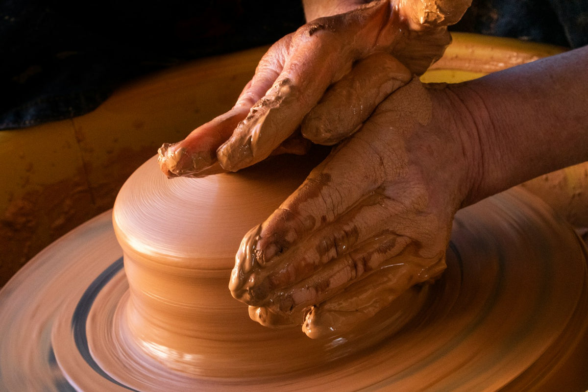 A potter's clay-covered hands shaping a pot on the wheel