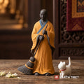 Sweeping monk statue in an ochre robe with the free white porcelain cat walking past him