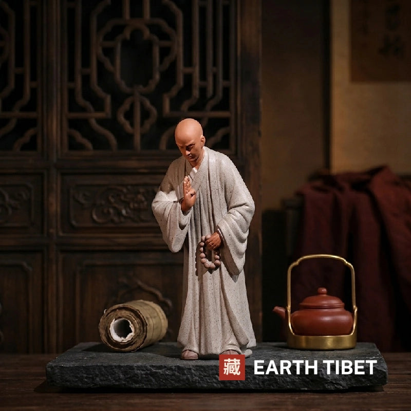 Grey ceramic zen monk statue with a hand raised in abhaya mudra beside a scroll and teapot