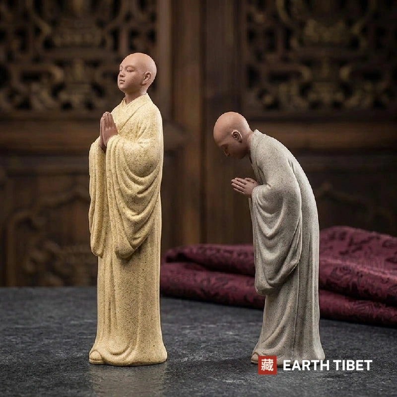 Beige standing monk statue with palms joined beside a grey monk bowed from the waist