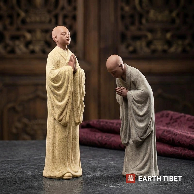 Two unglazed ceramic novice monk statues, one upright and one bowing, on dark stone