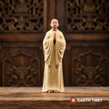 Front view of the beige standing monk statue, palms joined at the chest, bare feet below the robe